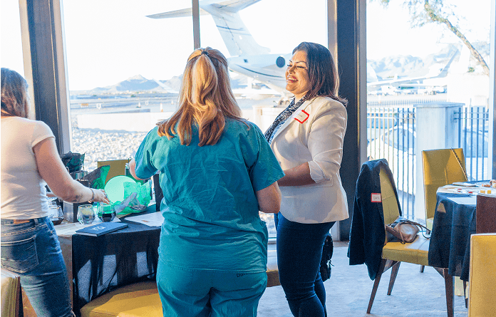 Three women at an indoor event with a view of an airport tarmac and private jet. A smiling woman in a white blazer talks to a woman in scrubs.