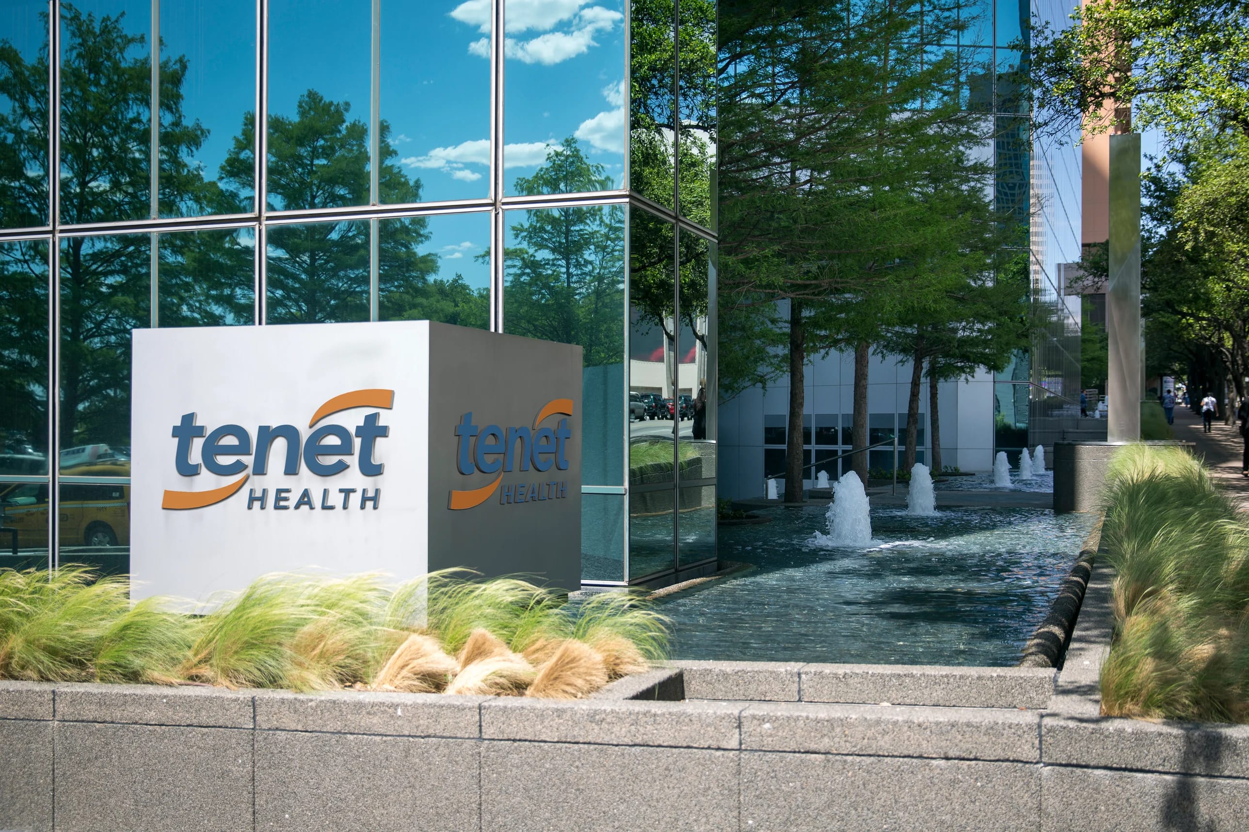The exterior of a modern building with blue reflective glass, featuring a Tenet Health sign in the foreground, and a water feature with several fountains surrounded by greenery.