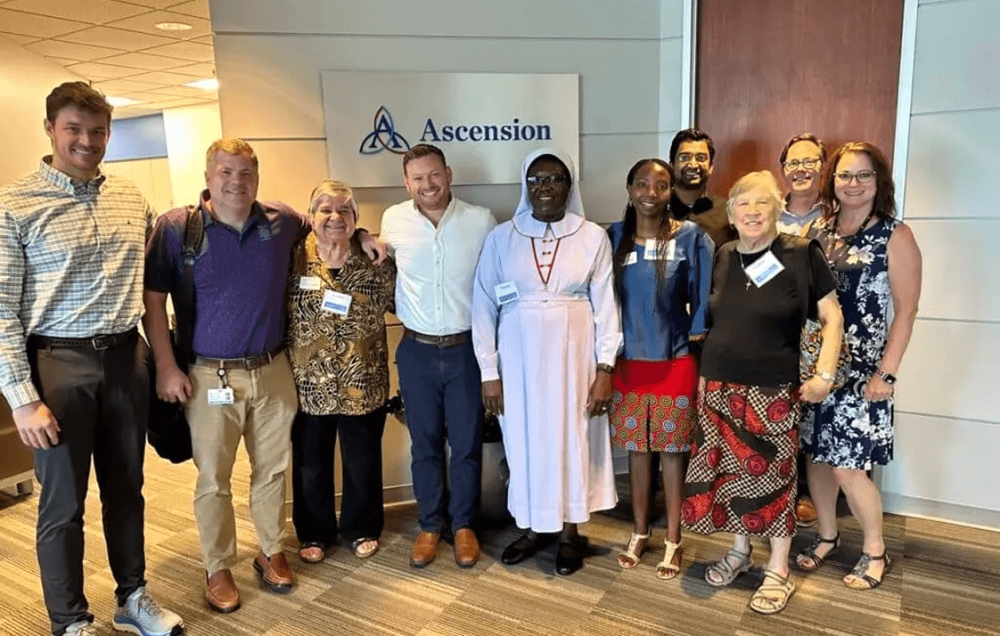 A diverse group of 11 smiling people, including a nun, pose together in front of an Ascension logo.