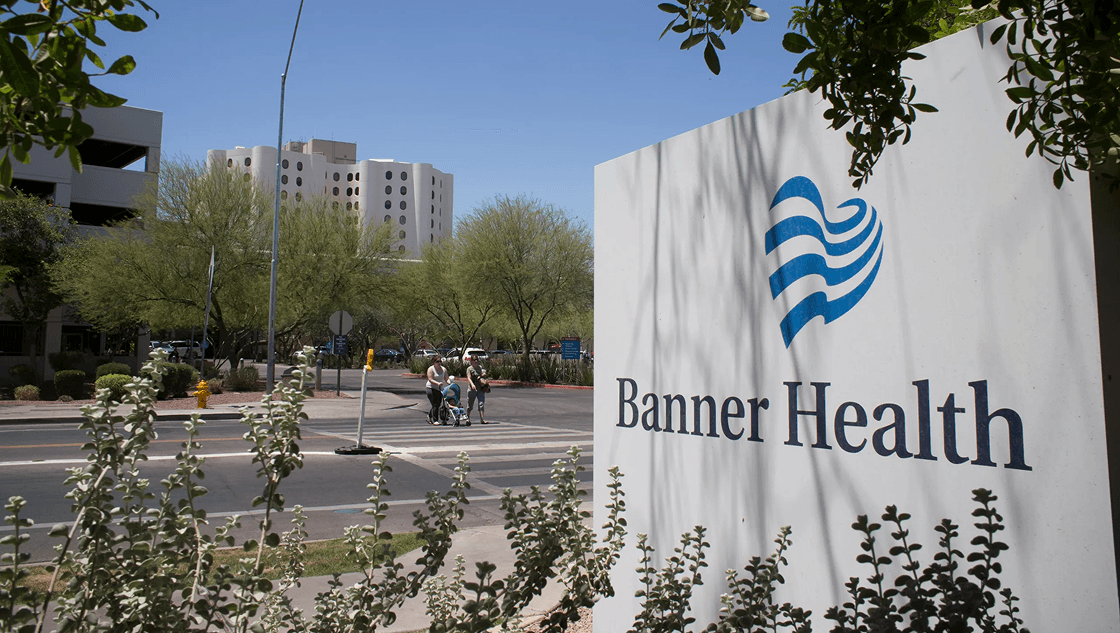 A prominent Banner Health sign in the foreground with a hospital building featuring round windows and people crossing a street under a sunny sky.