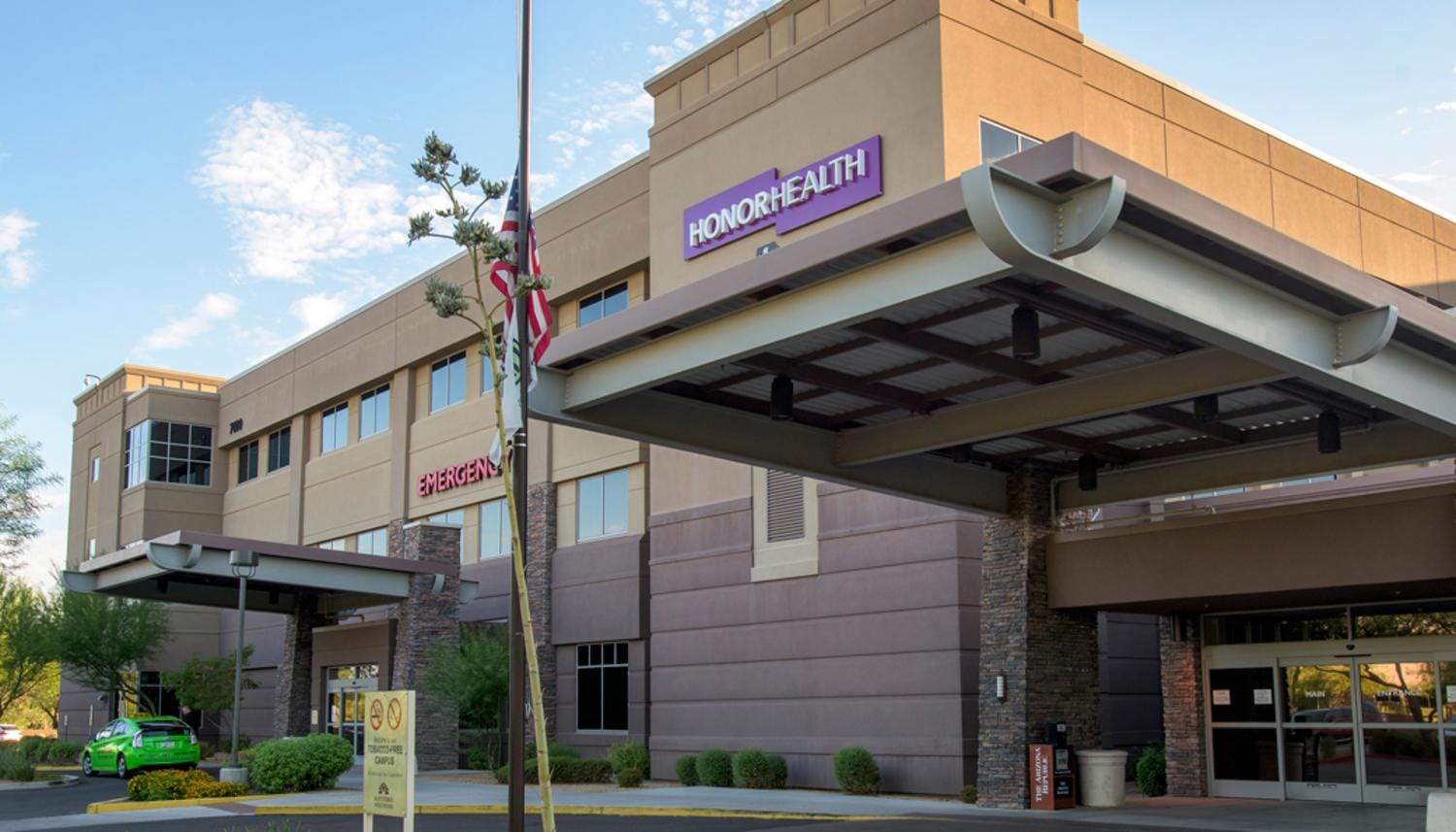 Exterior of the HonorHealth medical facility, featuring a beige building with purple "HONORHEALTH" signage, a clearly marked "EMERGENCY" entrance, and a covered main entrance area with flags.
